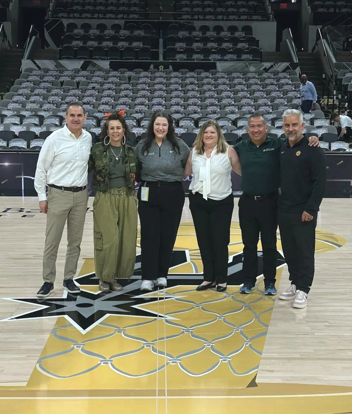 Group of six people standing on a basketball court with a large logo in the center, smiling and posing for the photo, with empty seating in the background.
