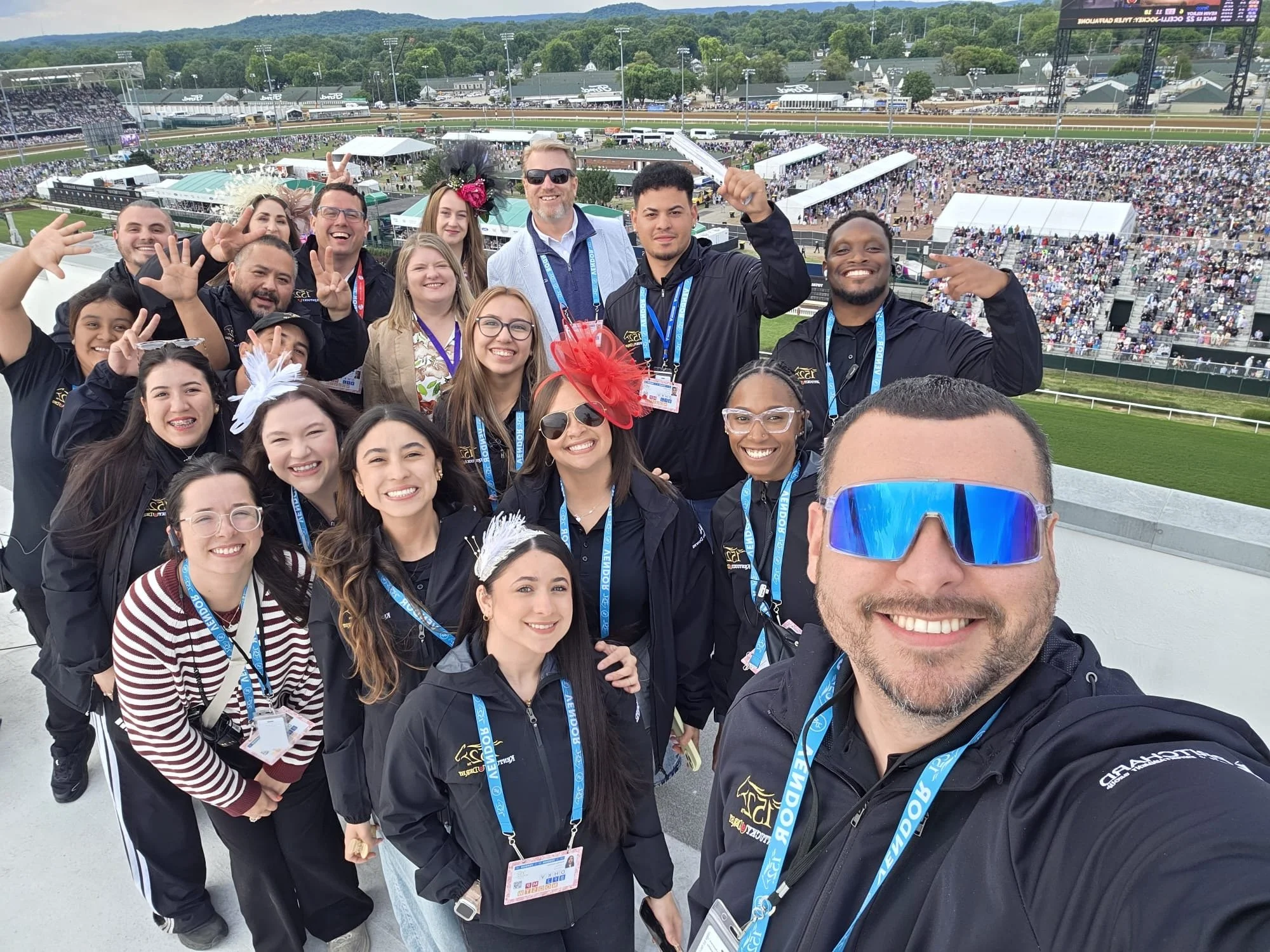 A group of people at a racetrack, smiling and posing for a selfie, with a large crowd in the stands and the racetrack in the background.
