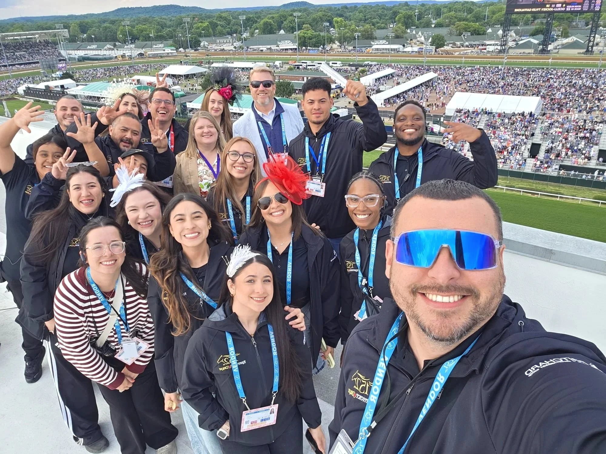 A group of diverse smiling people taking a selfie at a horse racing track with crowded stands and green hills in the background.