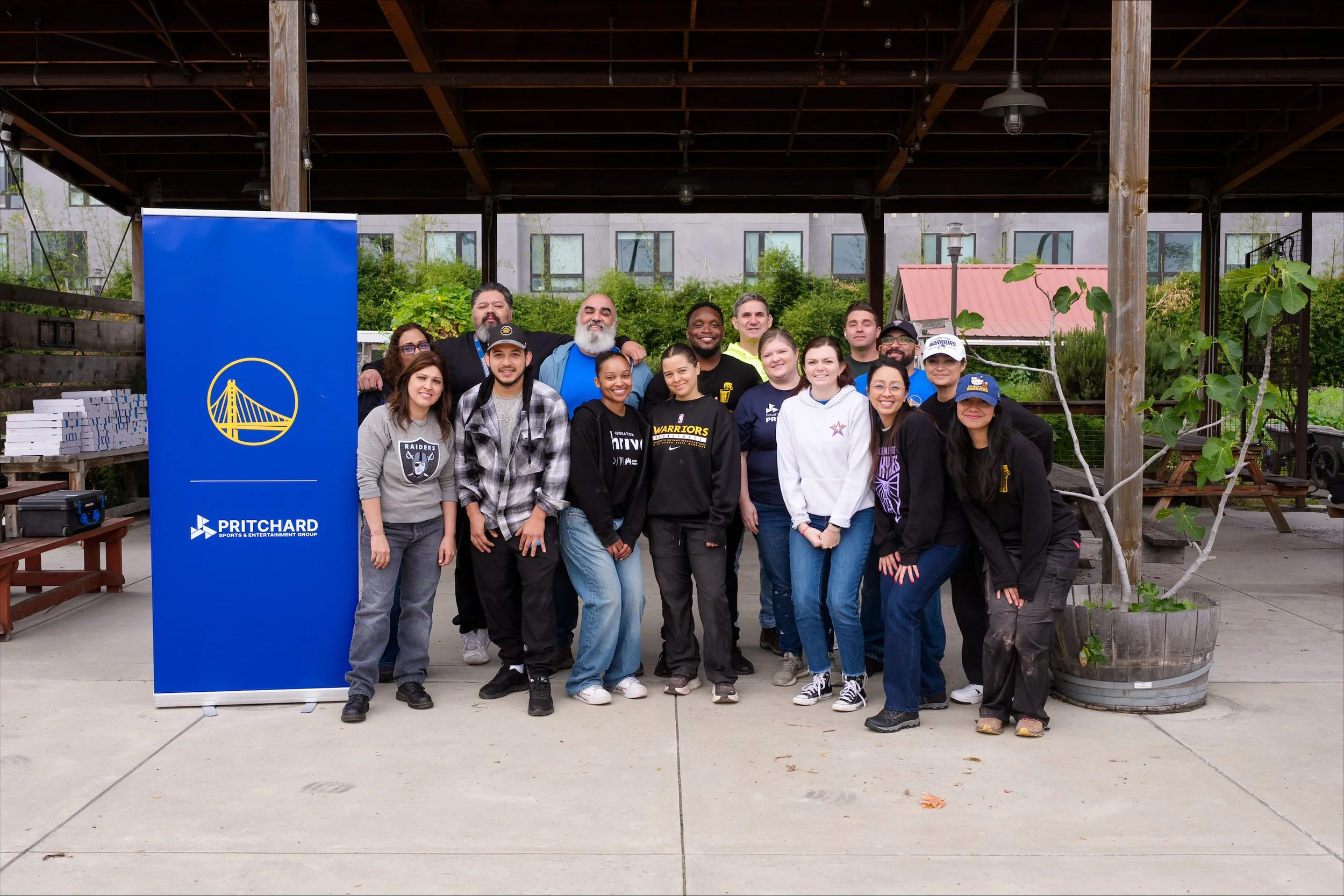 A group of diverse people posing together outdoors under a wooden pavilion, with a blue banner featuring a golden bridge logo and the words 'PRITCHARD' and 'SPORTS & ENTERTAINMENT GROUP' in front of them.