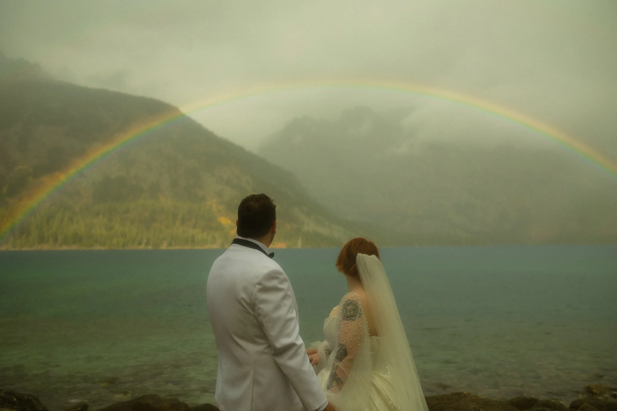 an elopement couple standing on a lakeshore with a rainbow in the background
