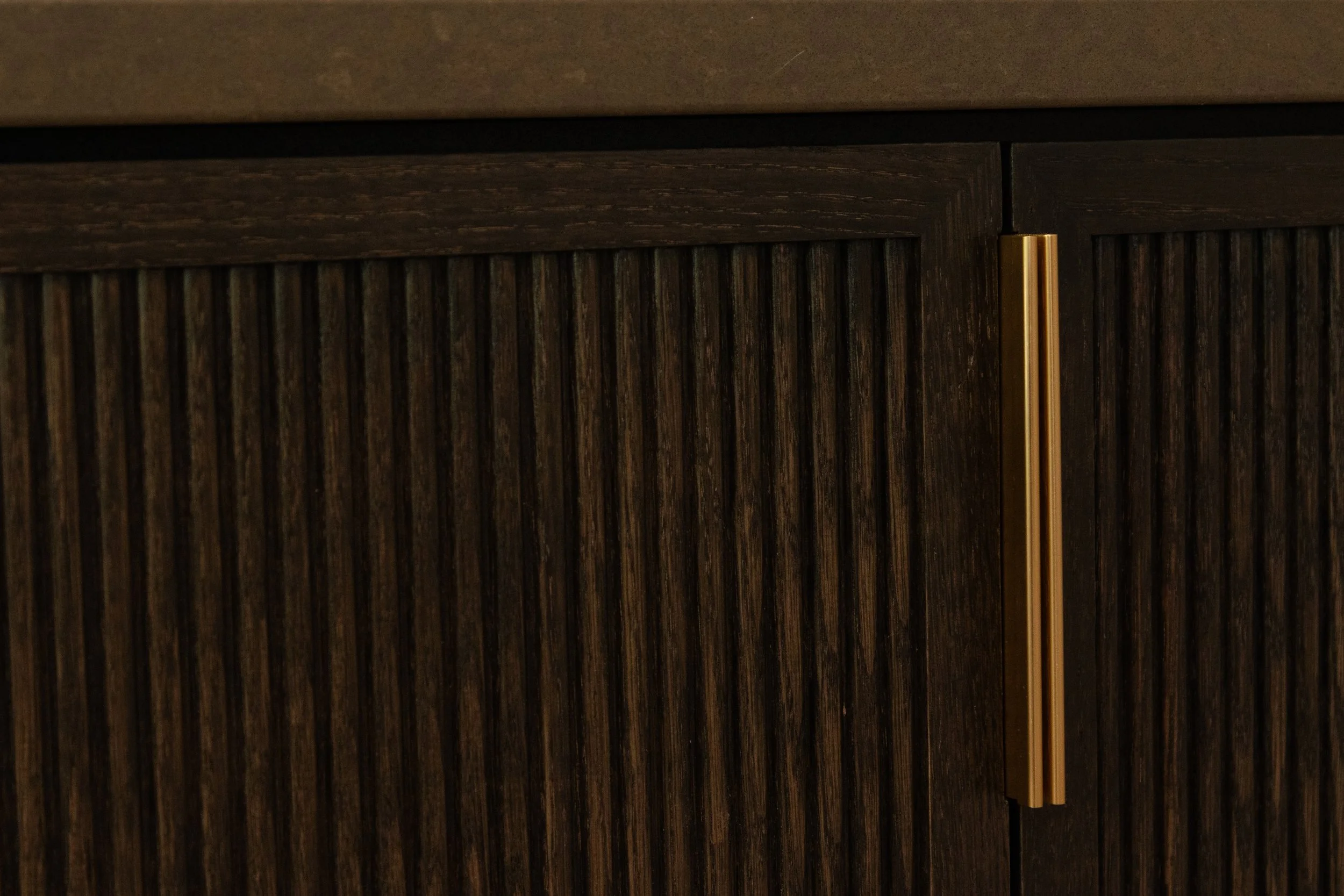 A dark wooden cabinet with vertical grooves and a gold handle, with a beige countertop above.