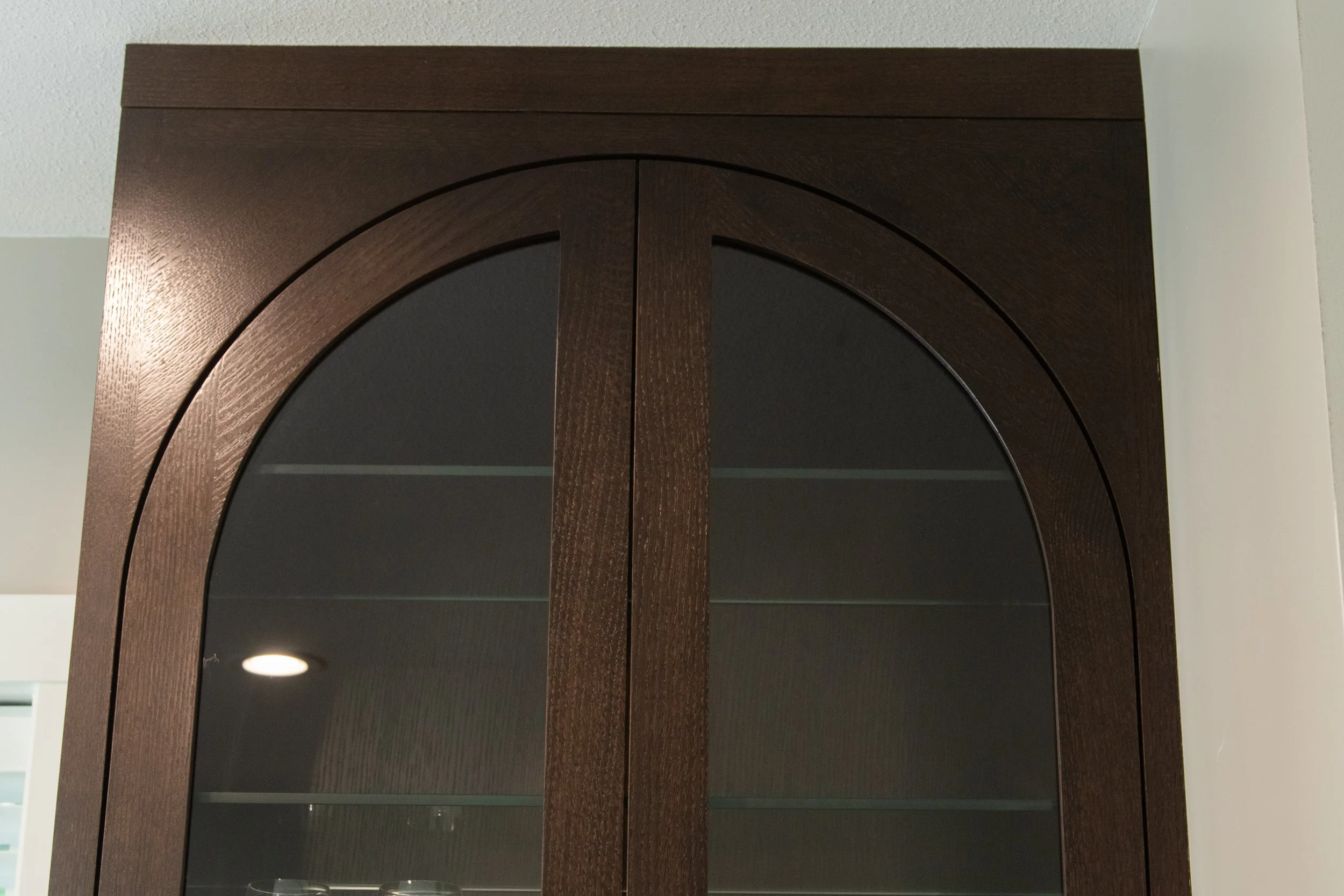 A dark stained oak cabinet with arched glass doors inset in an arched frame. with a white wall and ceiling in the background.