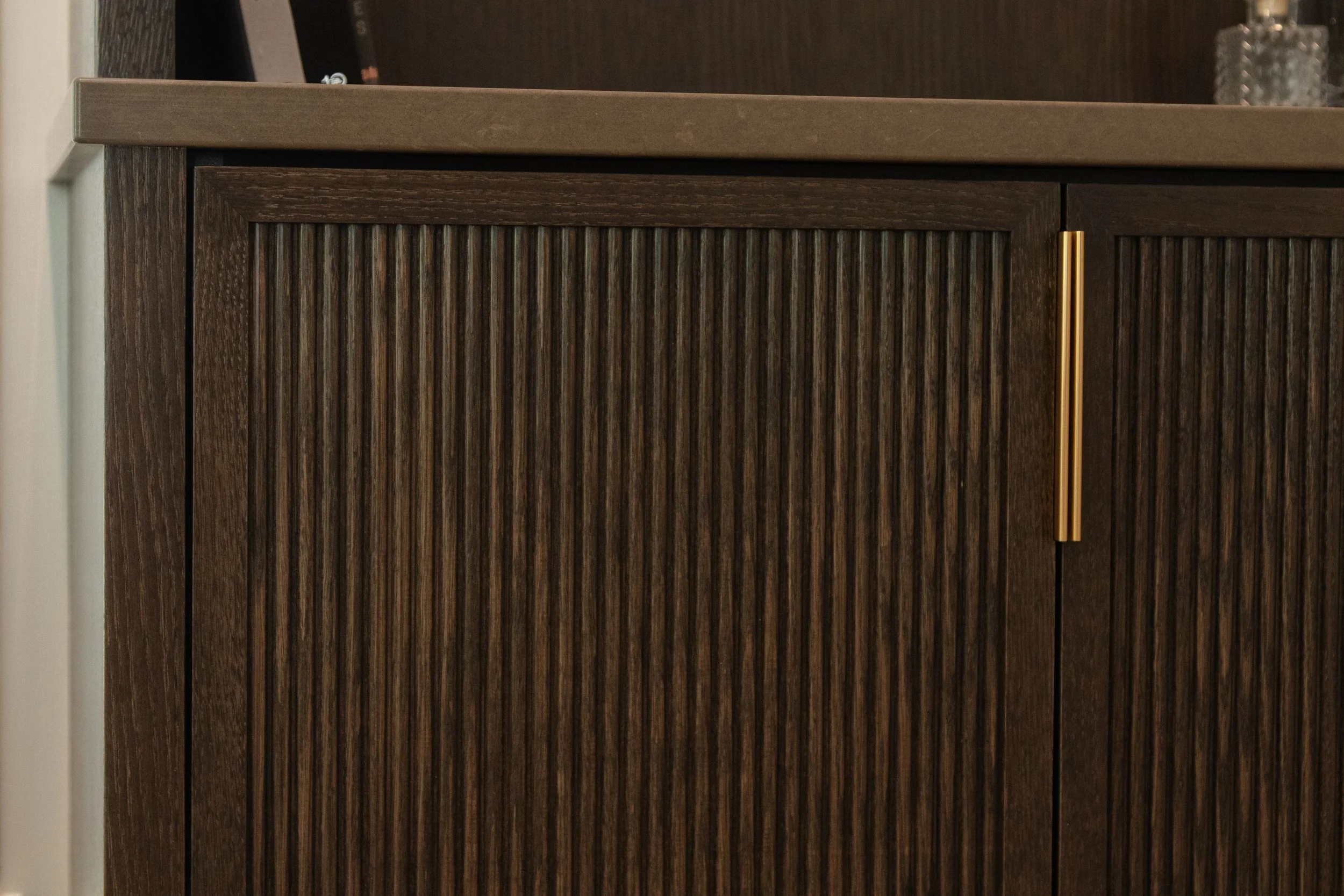 Close-up of a dark wood cabinet with vertical ribbed panels and a gold handle, topped with a dark brown shelf or table surface.