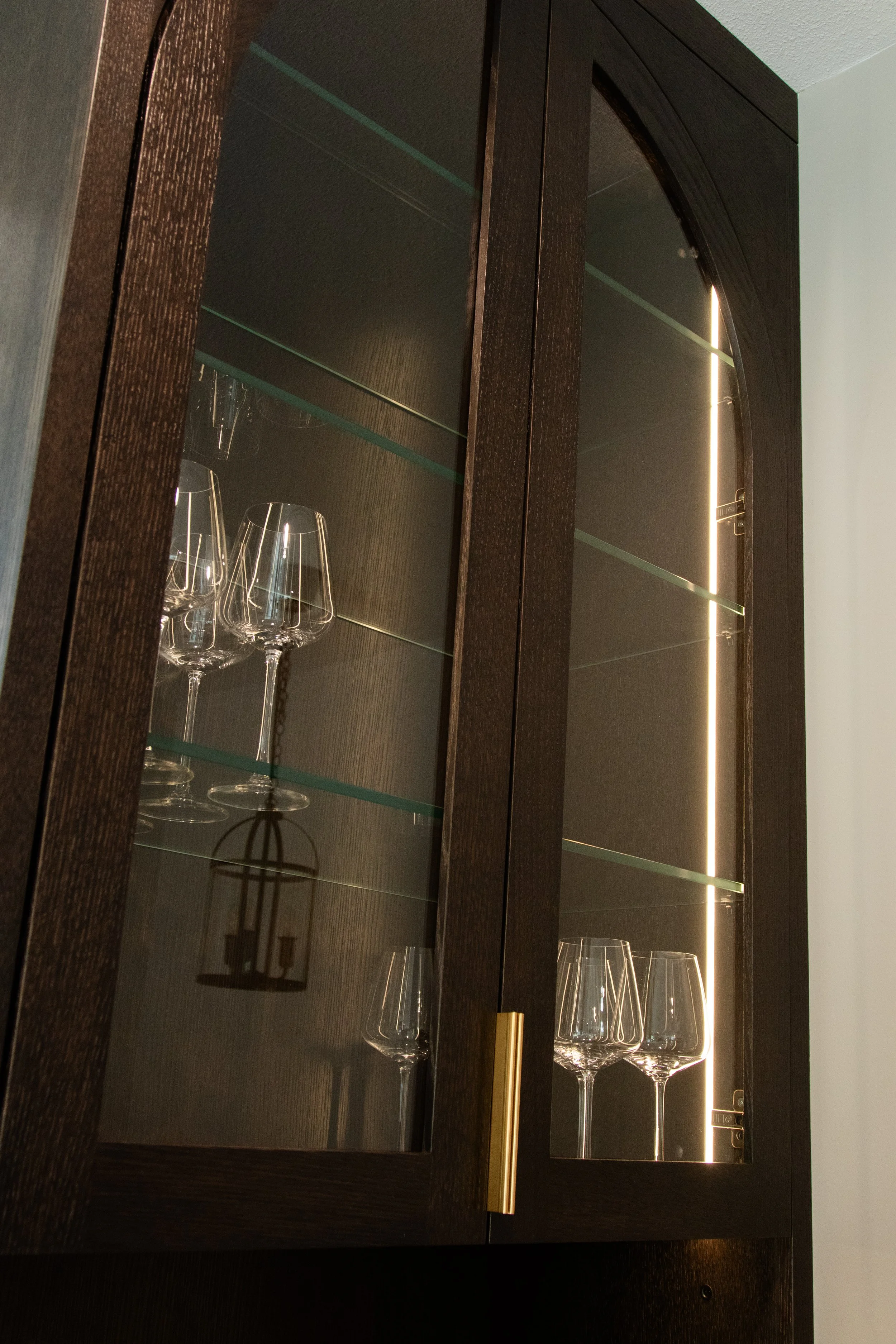A dark wooden cabinet with glass doors displaying empty wine glasses inside, illuminated by a vertical strip of LED lighting on the right side.
