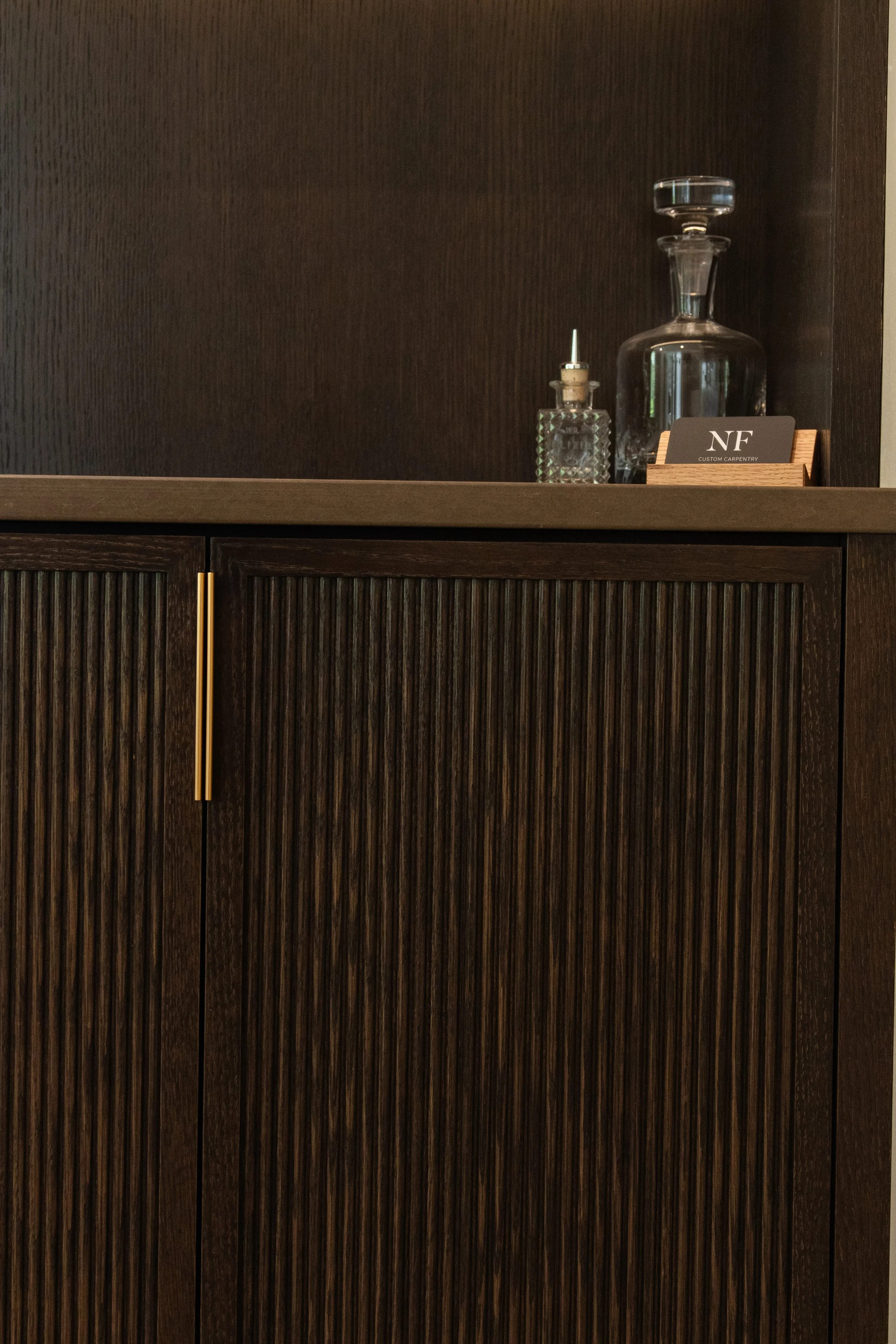 a Solid Oak wooden cabinet with vertical reeded doors doors, gold-colored edge pulls, and decorative glass bottles and business cards on top.