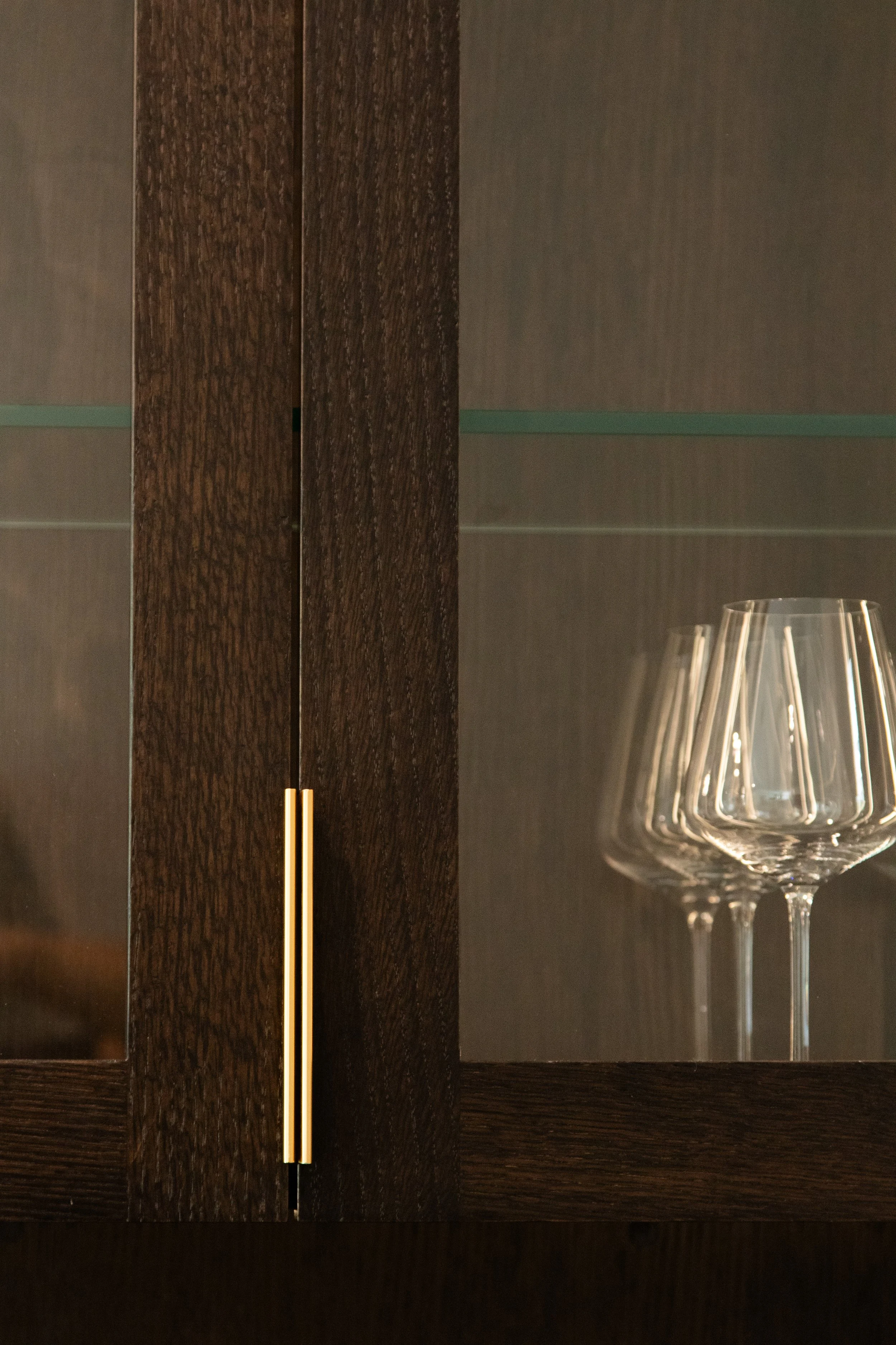 A wooden cabinet with glass doors, a vertical brass handle, and a set of empty wine glasses on a shelf inside.