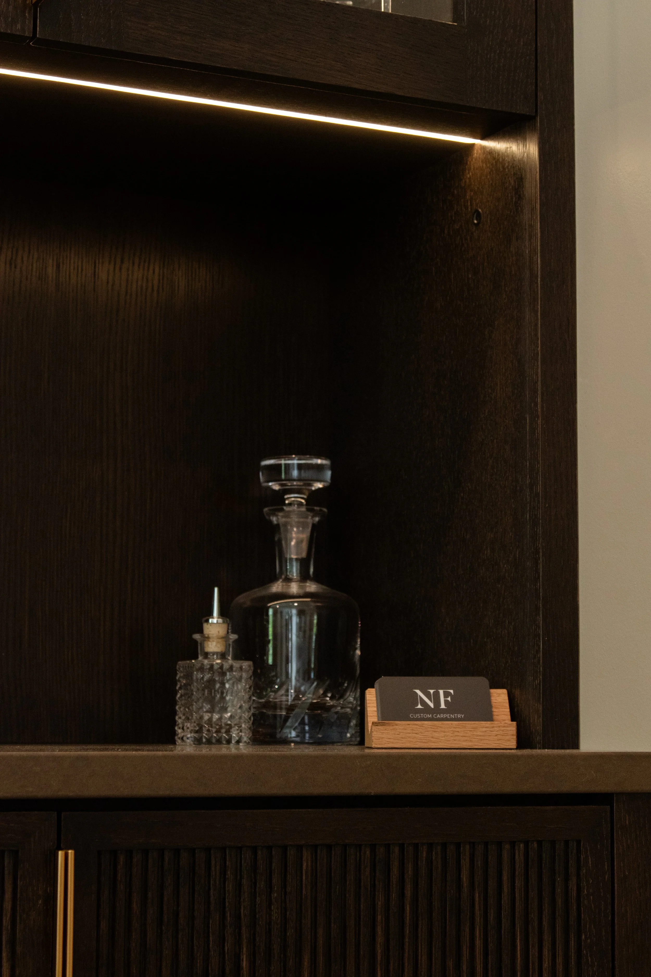 A dark wooden display shelf with under cabinet lighting holding various glass decanters, a small card with the initials at the bottom right, and a sign with the initials NF for Custom Carpentry.