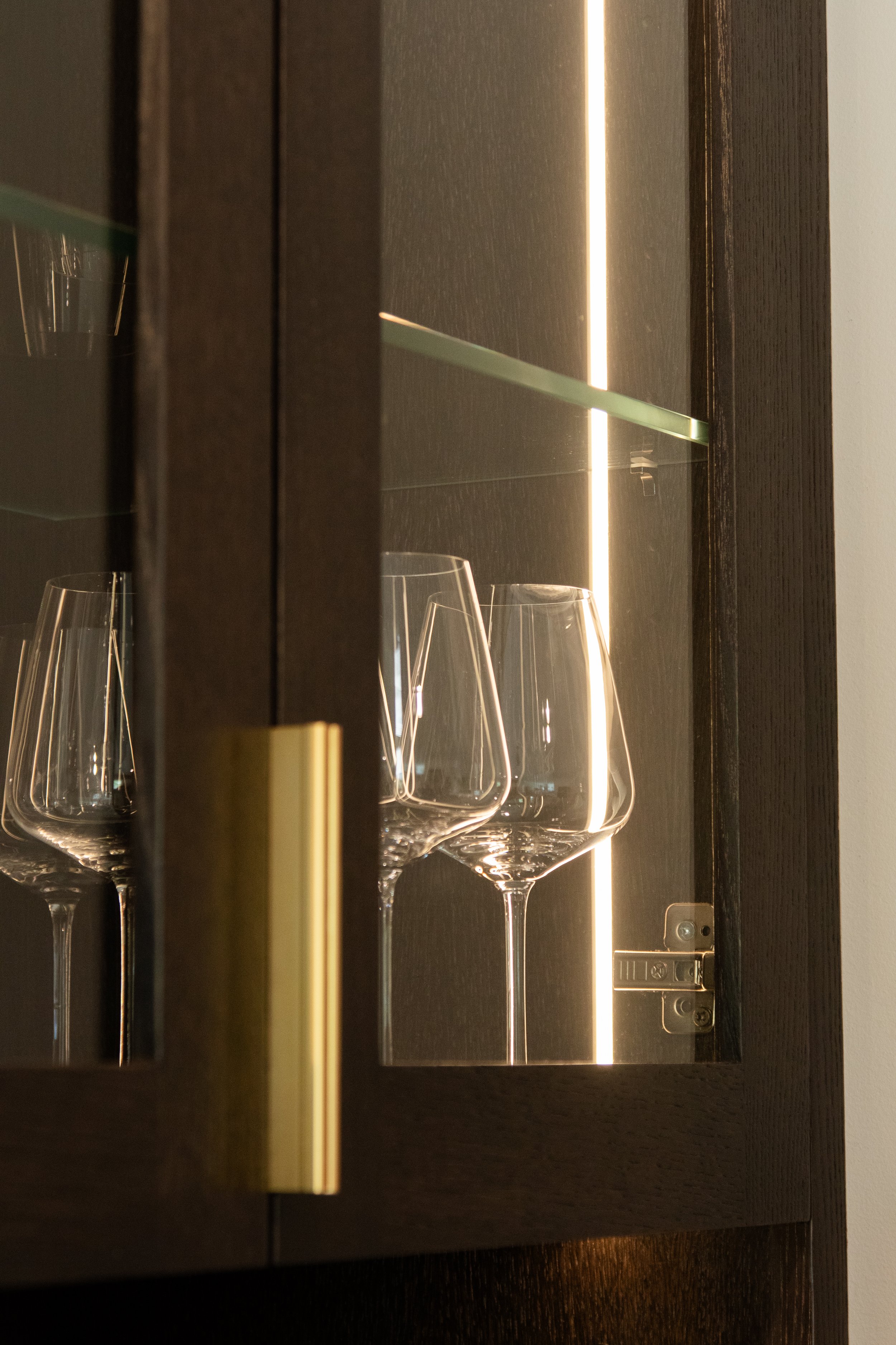 Four empty wine glasses arranged in a glass cabinet illuminated by a vertical LED light strip, with a dark wooden frame.