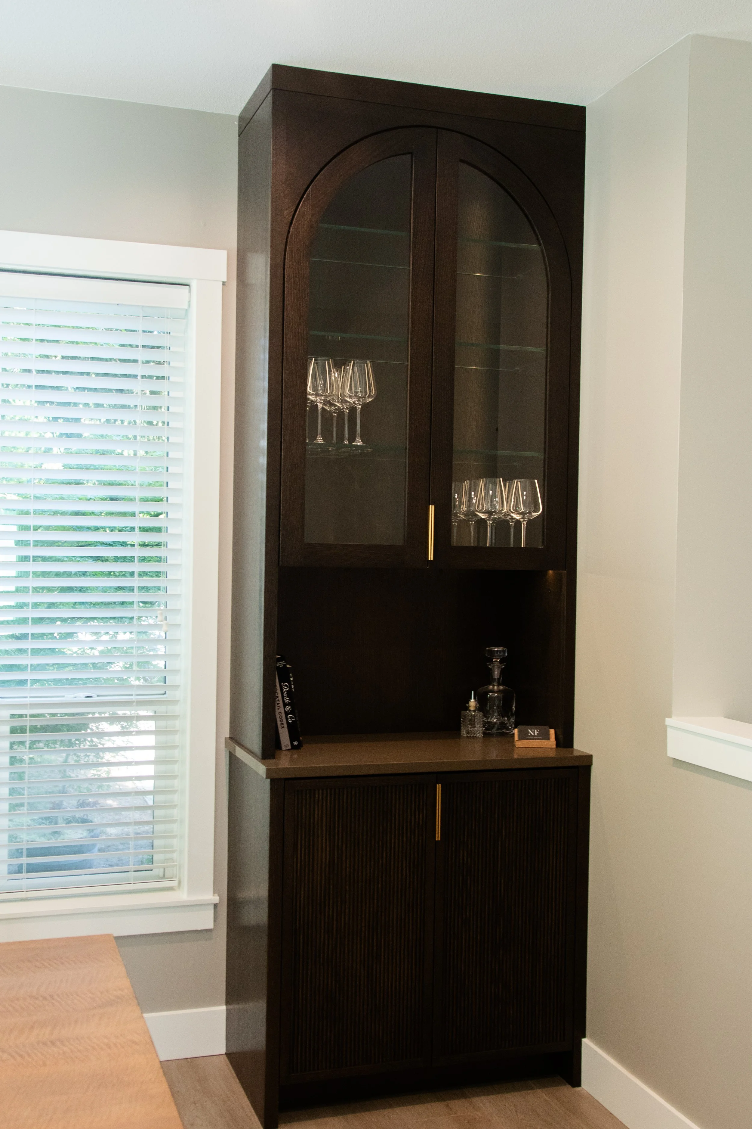 A dark wooden cabinet with glass doors containing wine glasses, situated in a corner next to a window with closed white blinds.