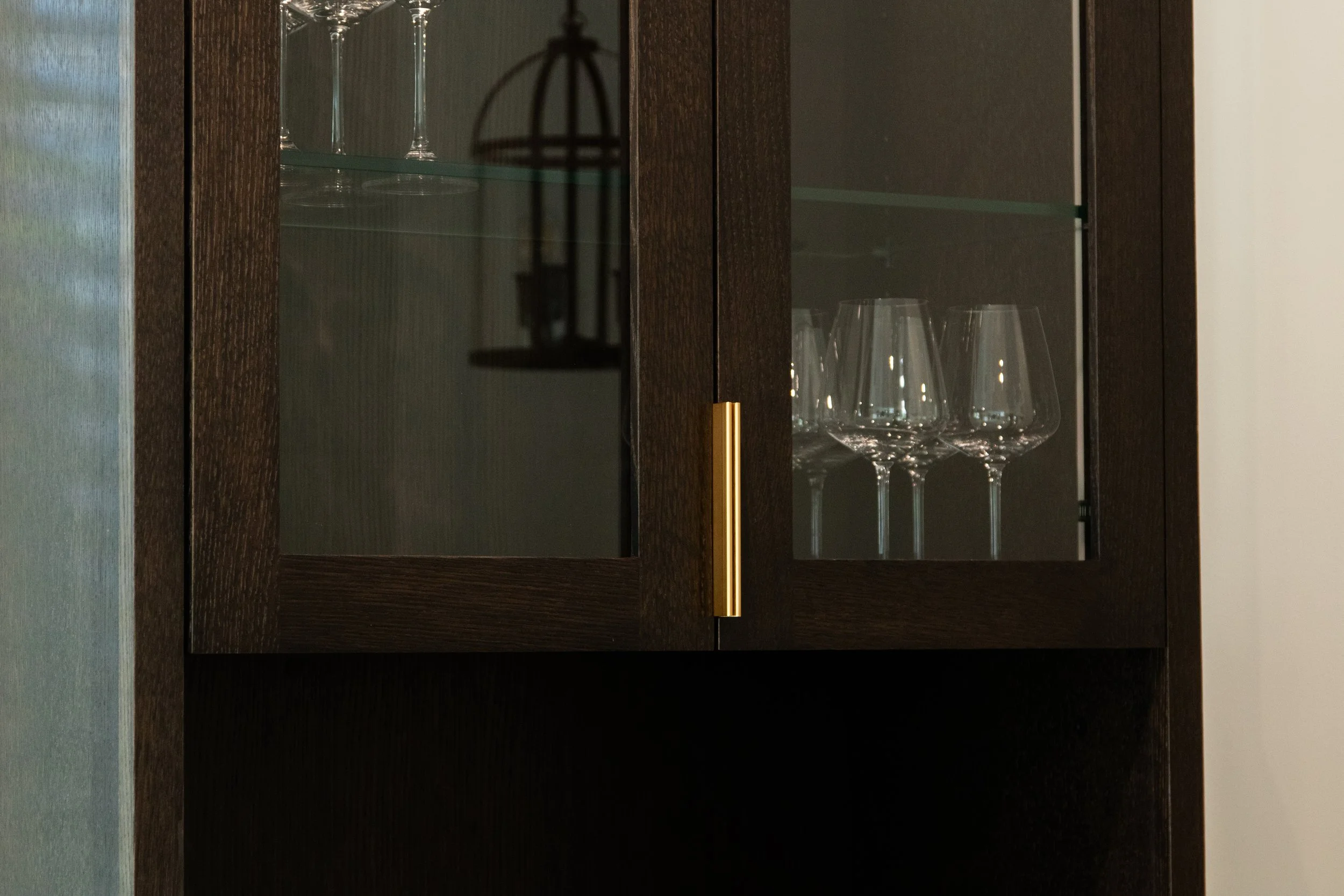 A dark stained oak cabinet with arched glass doors, containing wine glasses inside. The shadows of the glasses and a hanging birdcage are cast on the glass doors.