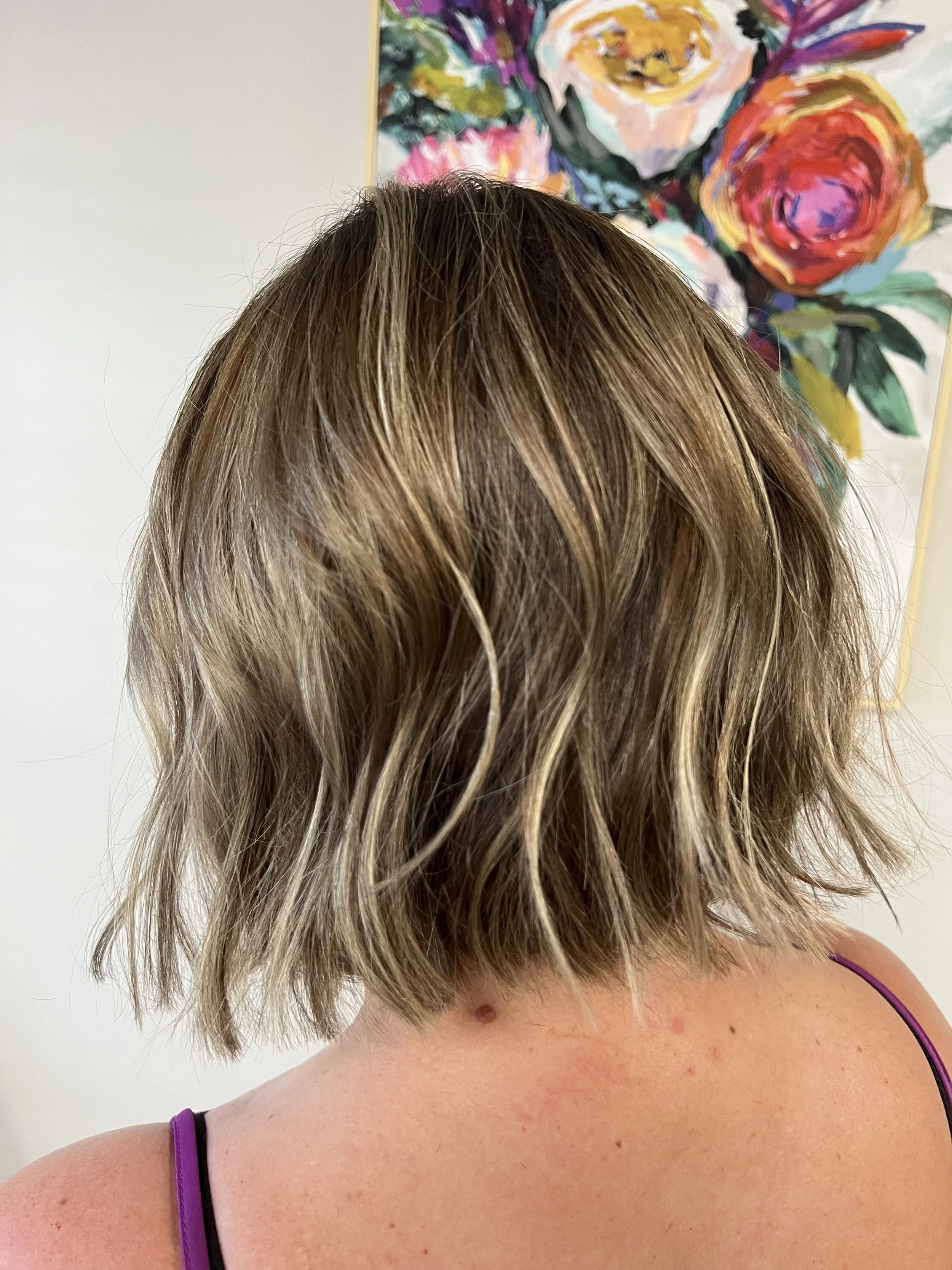 Close-up of the back of a woman's head with short, wavy hair that is a blend of brown and blonde highlights. She is wearing a purple strap top. In the background, there is a colorful abstract painting of flowers on a white wall.