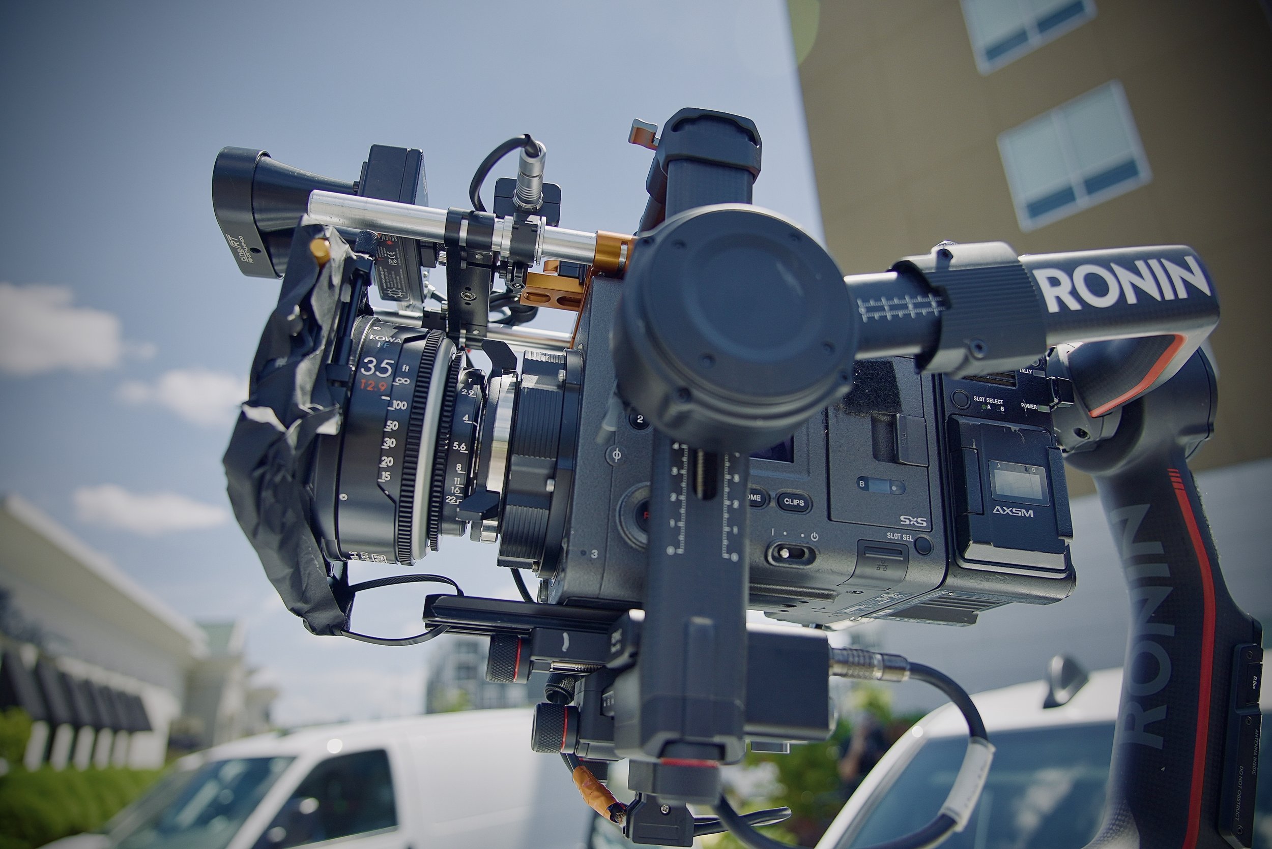 Close-up of a professional video camera mounted outdoors, with a building and sky in the background.