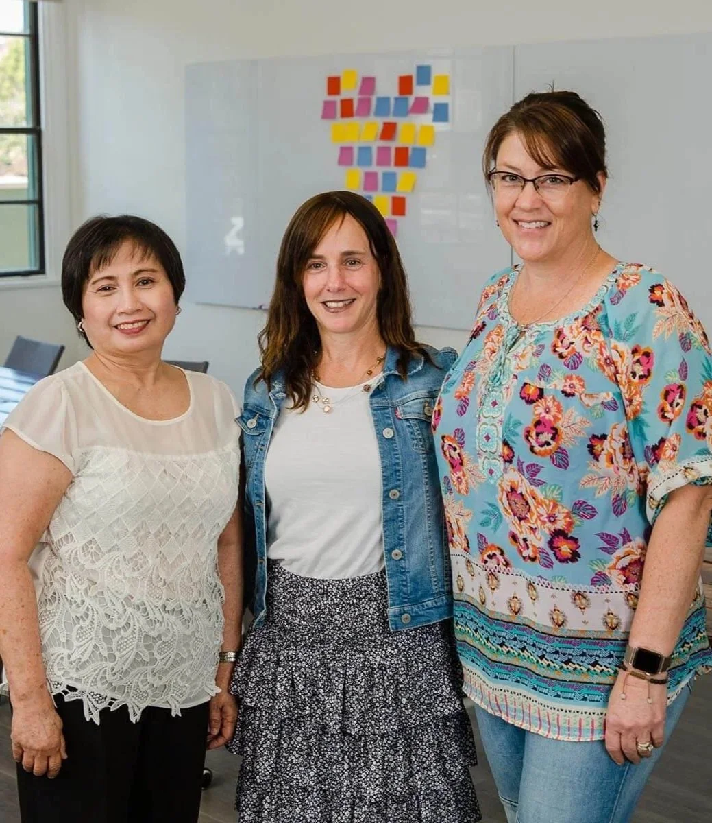 Three mission edge nonprofit accountants experts standing together in a brightly lit conference room with a large wooden table and a whiteboard with colorful sticky notes forming a heart in the background.