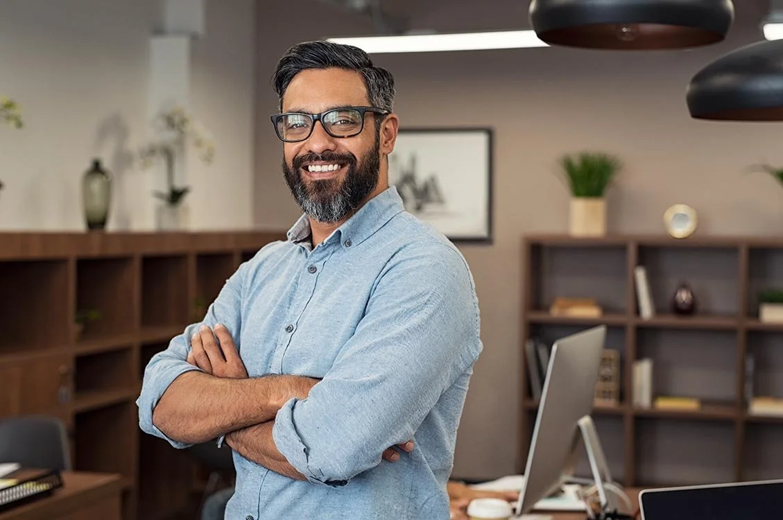 Smiling nonprofit manager standing in office with arms crossed and wearing a blue button down shirt.