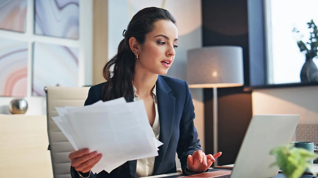 Nonprofit accountant wearing a blue blazer working at her desk typing on her computer and holding a stack of papers in her hand.