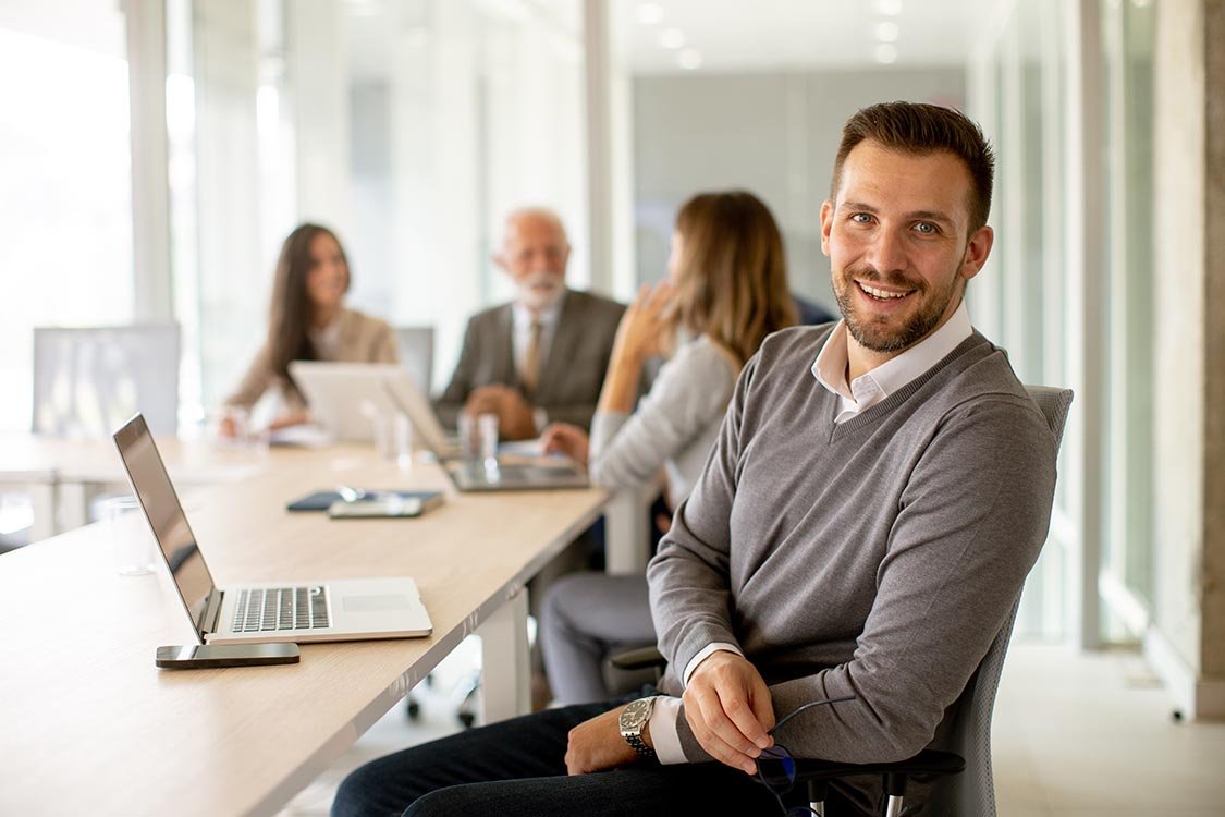 relaxed man leaning back in office chair in meeting space as people with laptops talk in the background