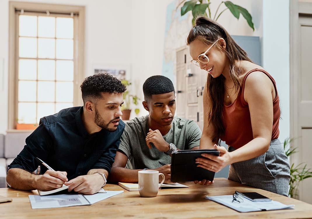 A woman stands at a table where her coworkers sit and shows them what she's been working on on her tablet as one of them takes notes.