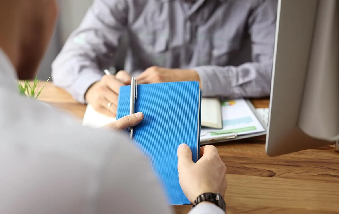 Employee talking to manager holding blue handbook with silver pen.