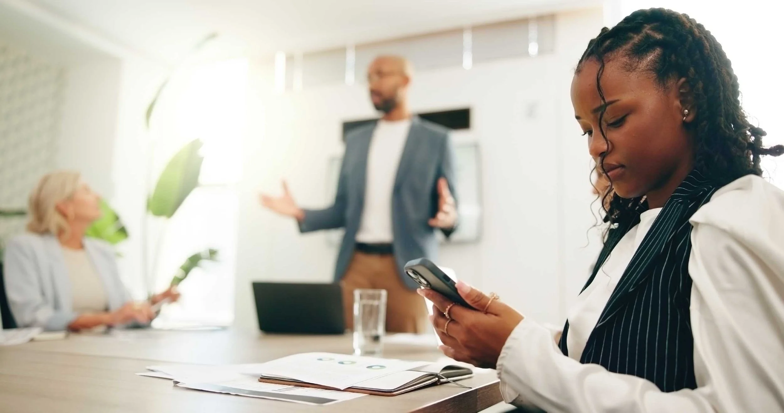 Young nonprofit employee checking phone and disengaging from meeting while nonprofit management is talking.