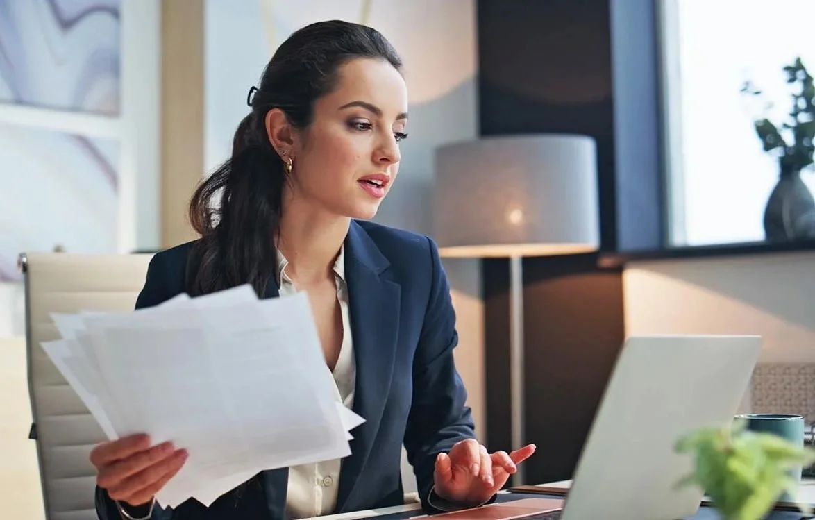 nonprofit accountant wearing a blue blazer working at her desk typing on her computer and holding a stack of papers in her hand.jpg