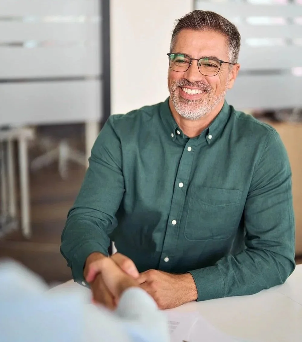 confident middle aged man with glasses smiling as he shakes hands at the beginning of an interview.