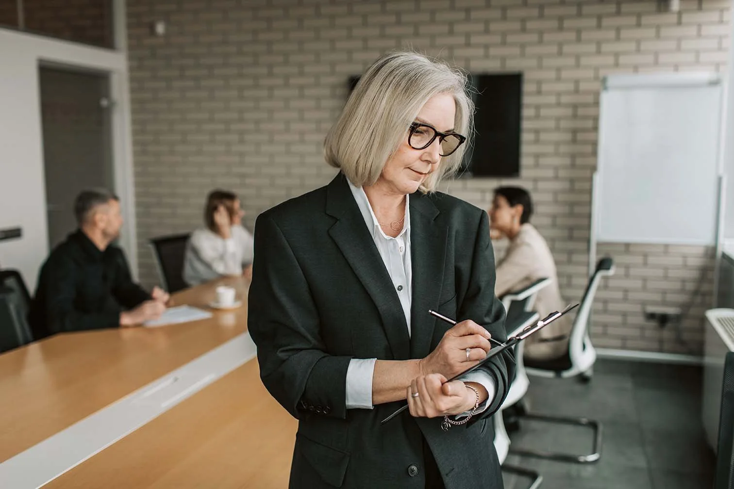 Nonprofit board member standing and taking notes on a clipboard while the board sits at a long table and discusses hiring someone.