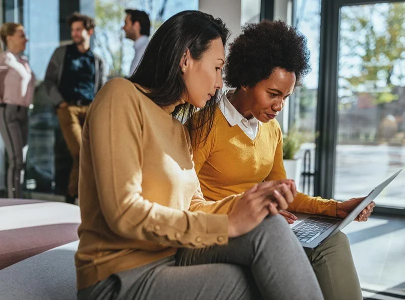 Two women sitting in office near windows intently reviewing something on a laptop together.