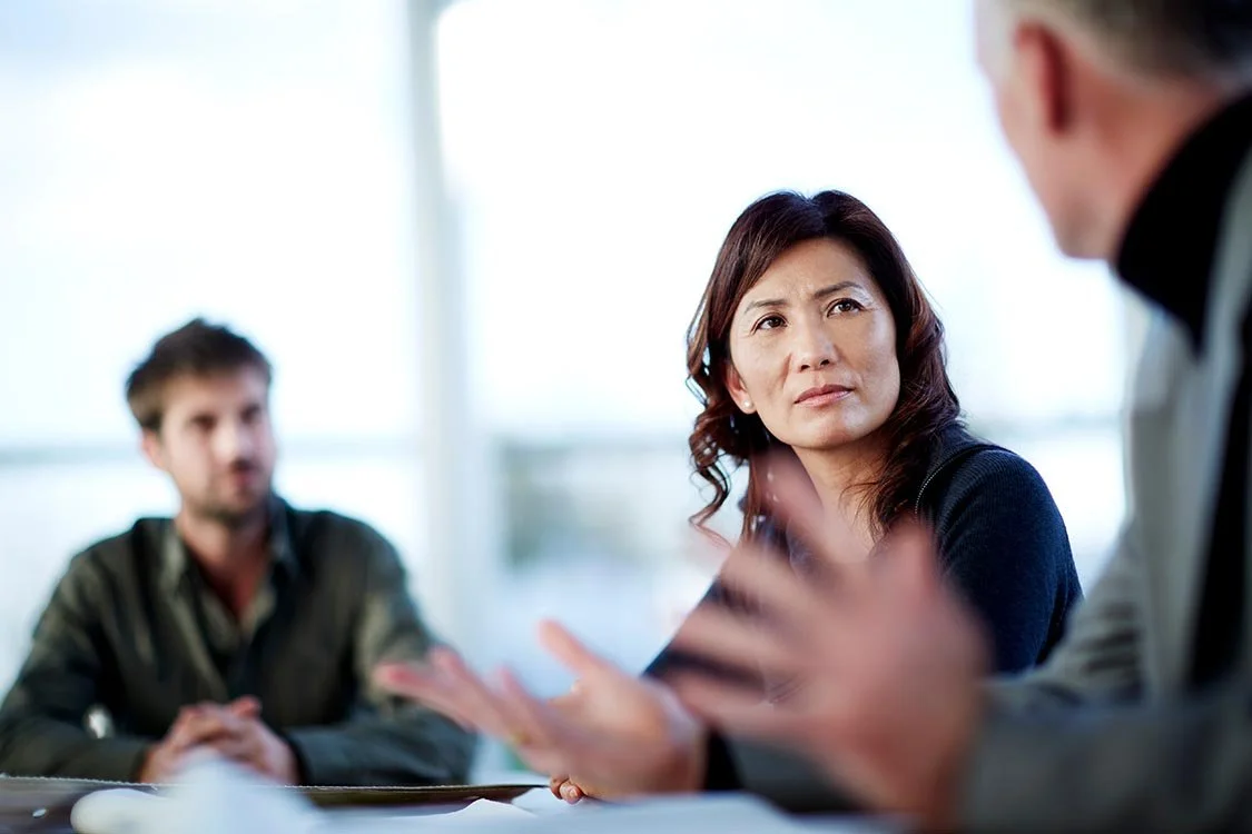 Man with papers in front of him gesturing while he talks with two employees intently listening to him.