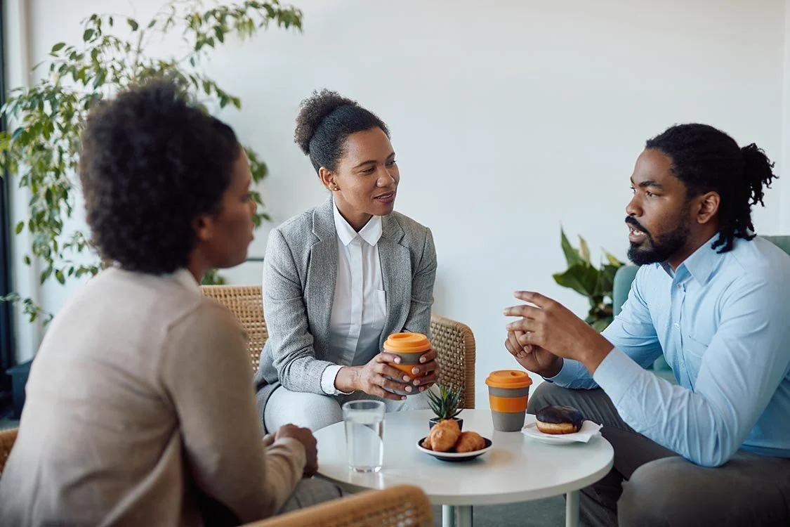 Nonprofit executive in blue button-up shirt talks with two women wearing blazers while sipping coffee in a cafe.