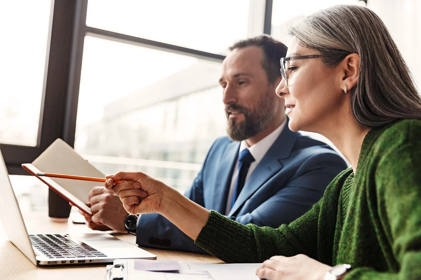 Woman wearing green sweater and man in blue suit review information together on laptop while taking notes.