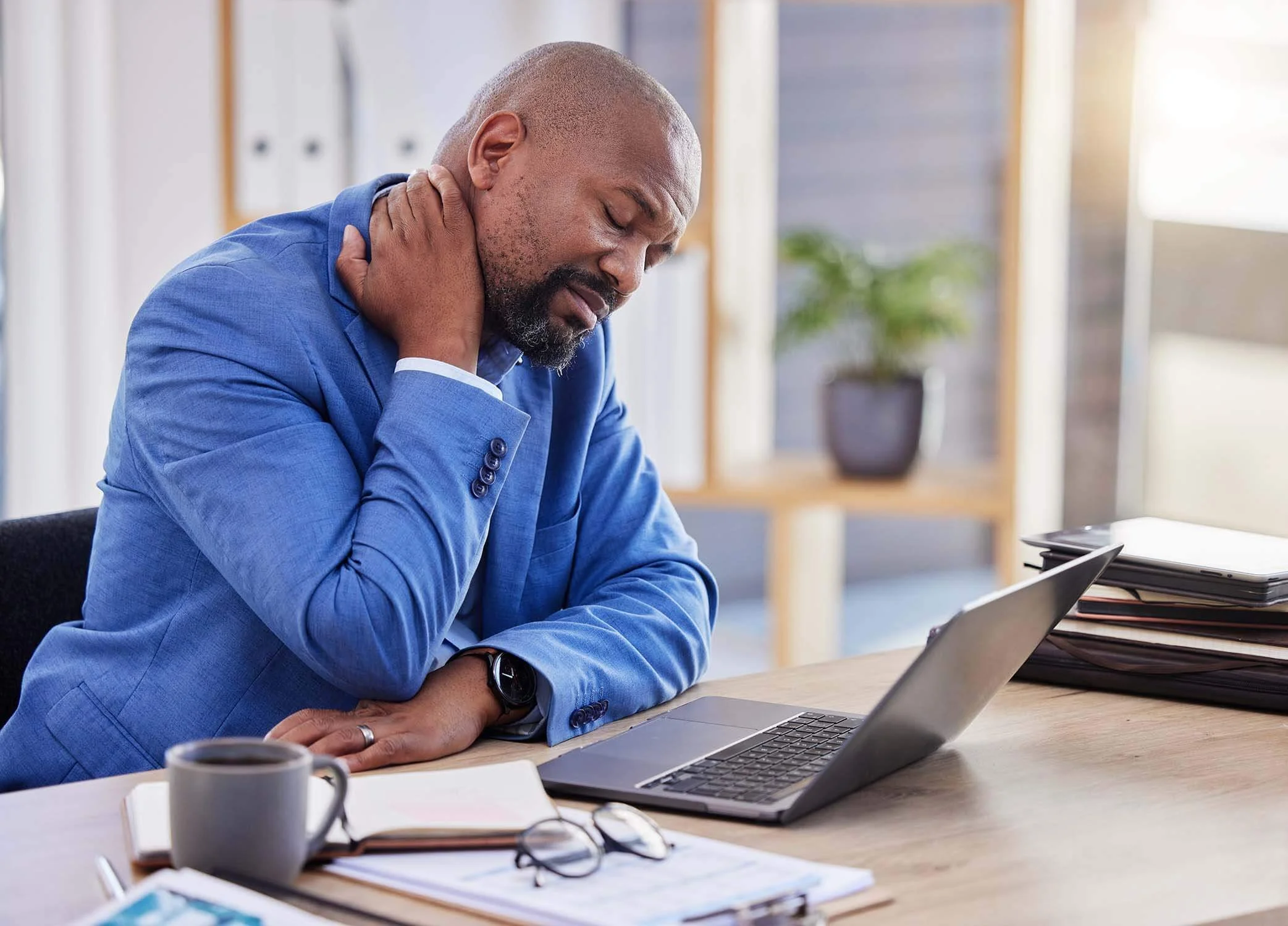 Nonprofit CEO stressed while working at his desk and rubbing his neck while he reviews documents on his laptop.