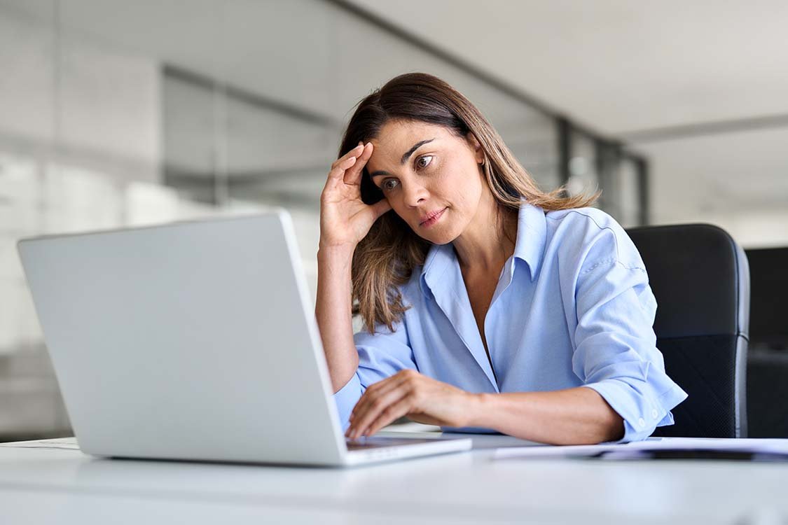 Woman wearing professional clothes looking frustrated and tired while working on her white laptop.