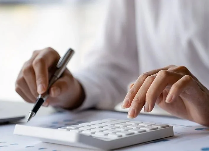 Person typing on calculator with one hand and making notes on their accounting reports with the other.