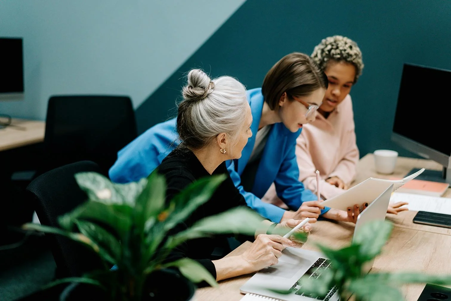 three women intently reviewing paperwork together at desk with multiple computers on it