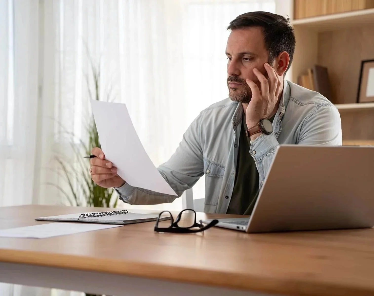 Nonprofit leader sitting at his desk with a bookcase behind him is focused on reviewing the printed accounting report printed in his hand while his open laptop sits next to him.
