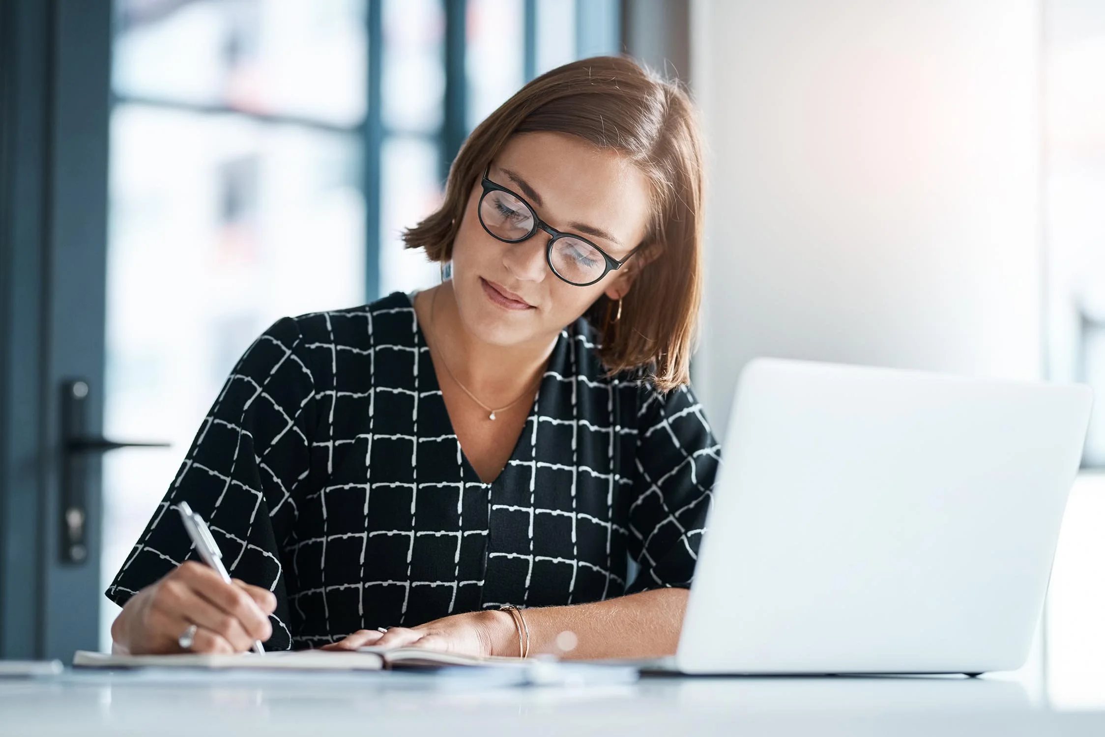 Nonprofit grants manager writing down ideas in notebook while working on her computer.