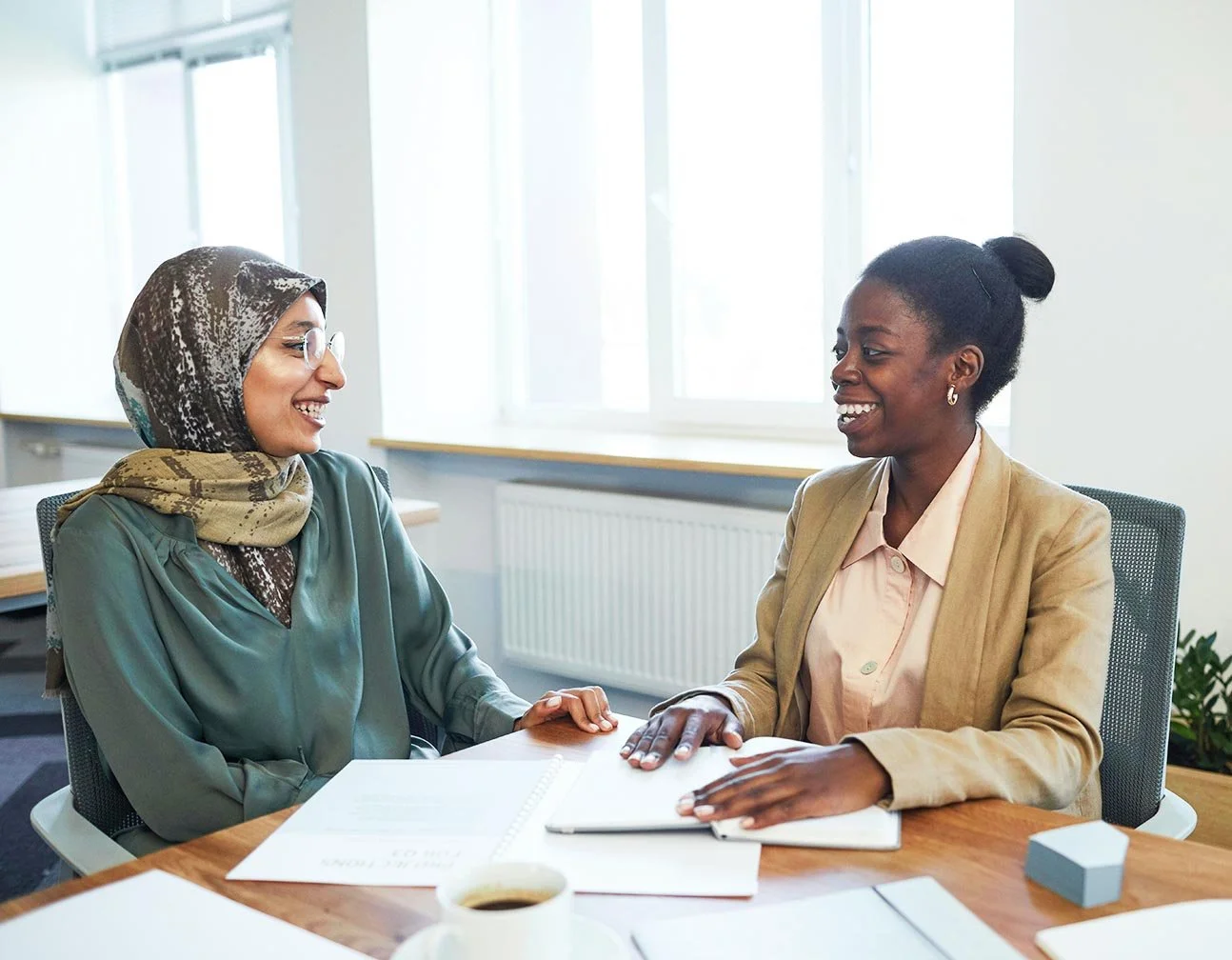 Two women, one wearing a headscarf and the other a blazer, smile and talk at a desk with an open hr performance report between them