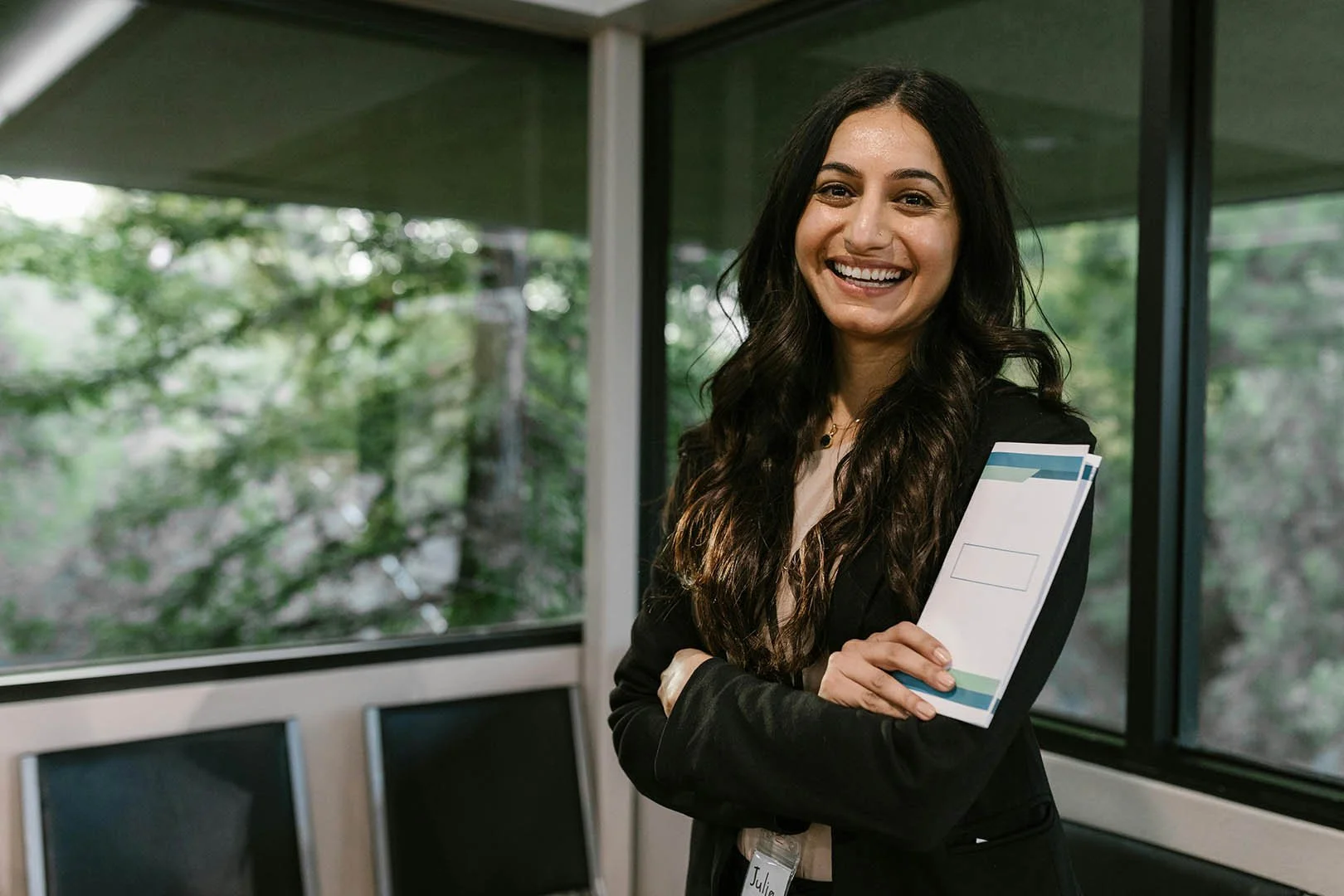 Smiling professional standing confidently indoors near large windows, holding a folder and wearing a name badge