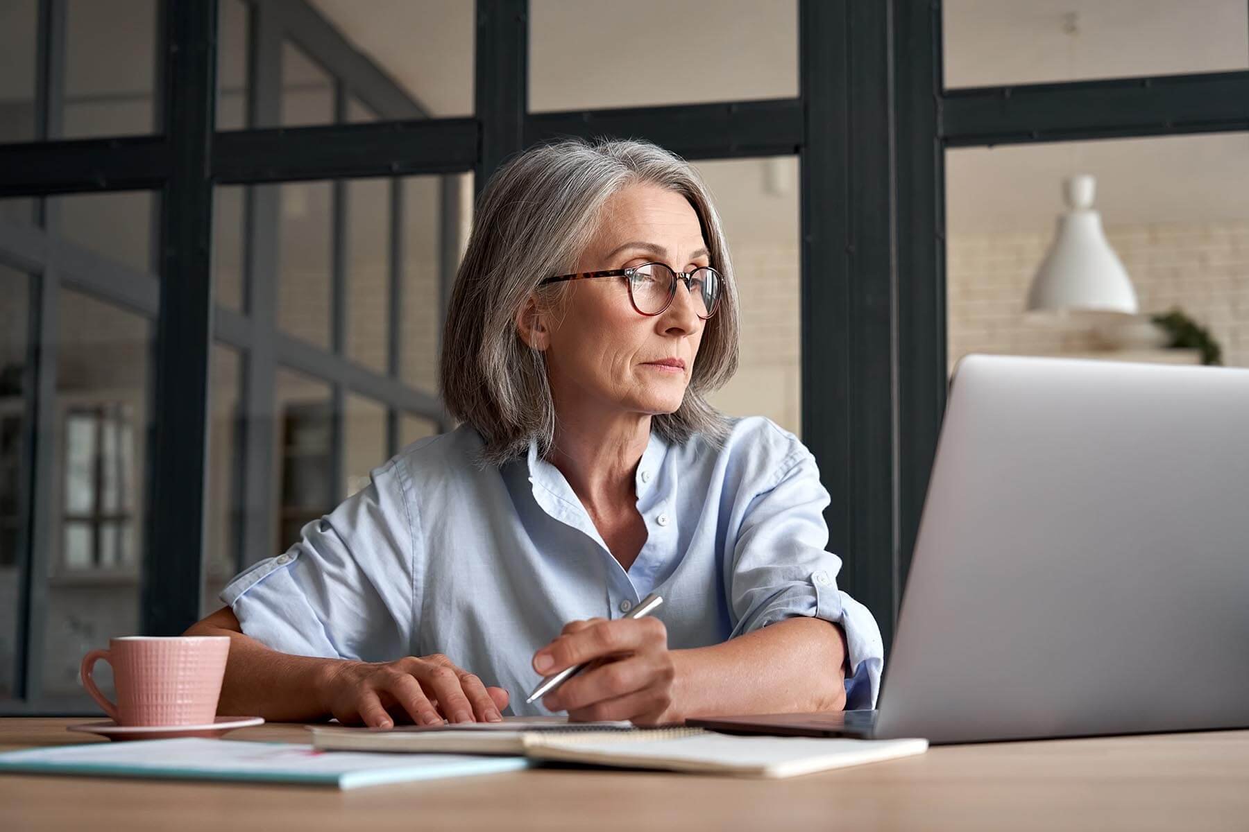 Older woman with gray hair and wearing glasses carefully reviews grant applications on her laptop while taking notes with a silver pen.