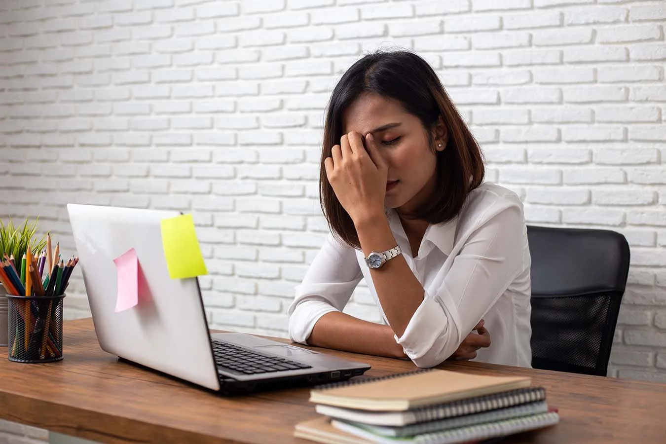 exhausted woman sitting at desk with her hand in front of her face in front of laptop with sticky notes on it
