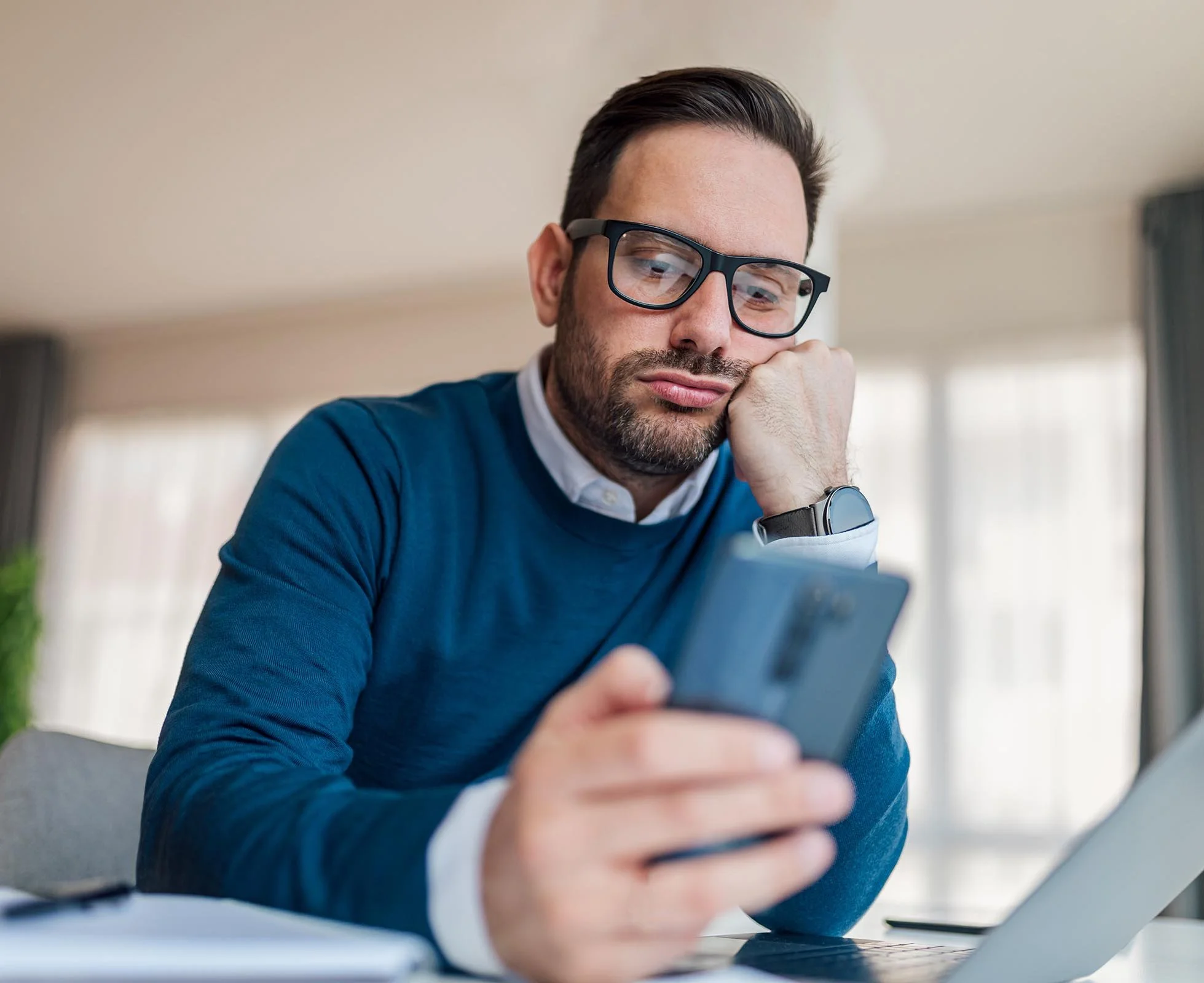 bored nonprofit employee looking at his smart phone instead of his laptop when sitting at desk.