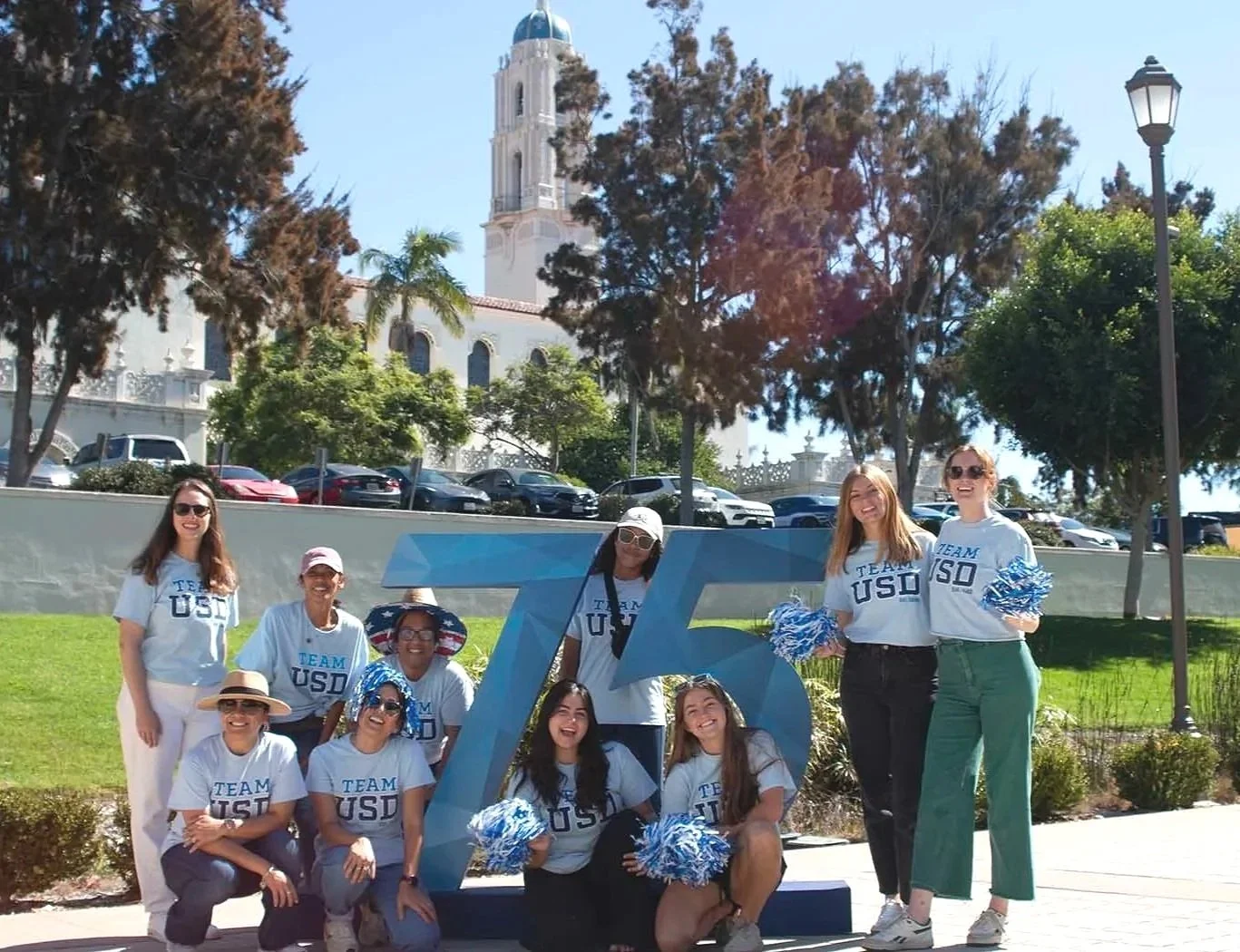 group of university of san diego volunteers holding pom poms and wearing matching team usd shirts while standing around 75 sign