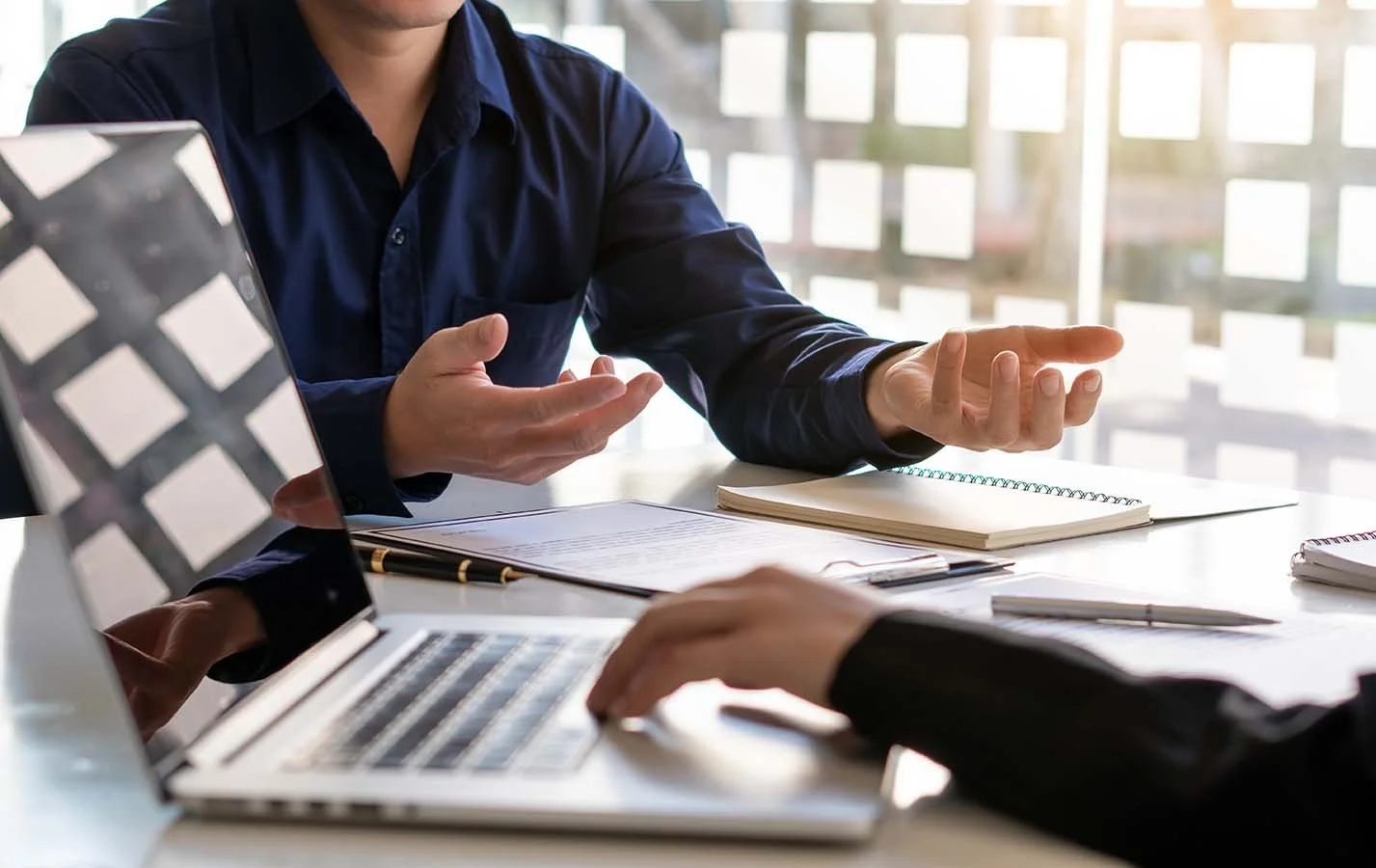 nonprofit leader with blue button-up shirt gestures with hands while talking to consultant at desk taking notes on laptop