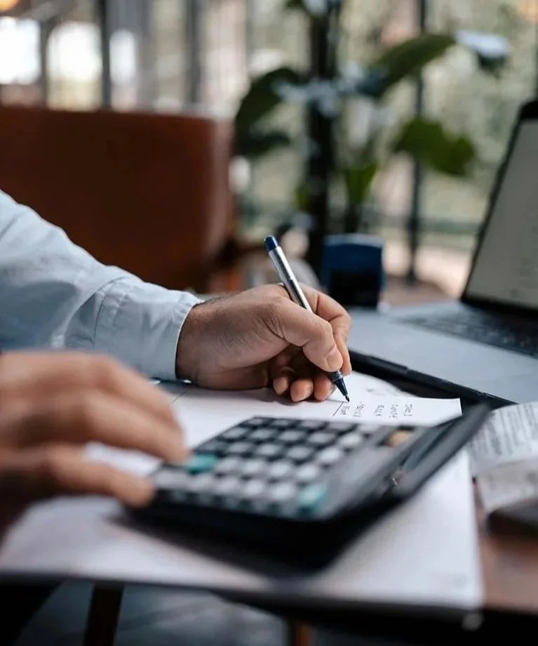 man sitting at desk next to large window writing down numbers while typing on calculator
