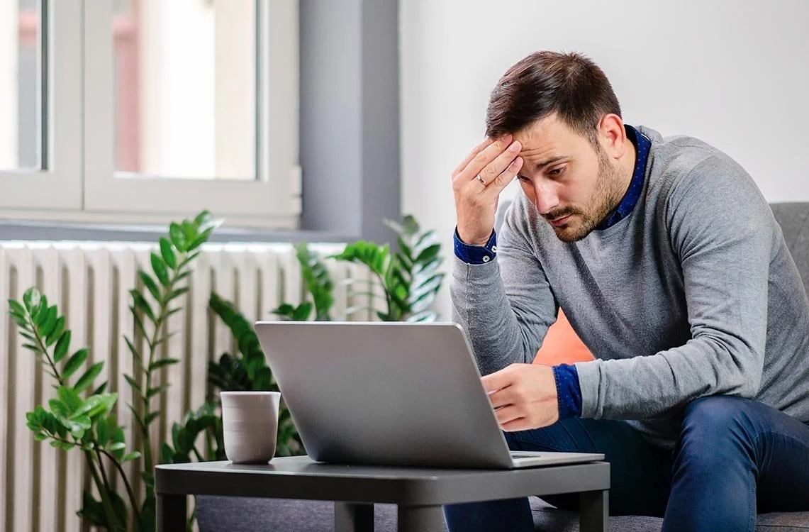 tired man in grey sweater working on laptop and rubbing his forehead