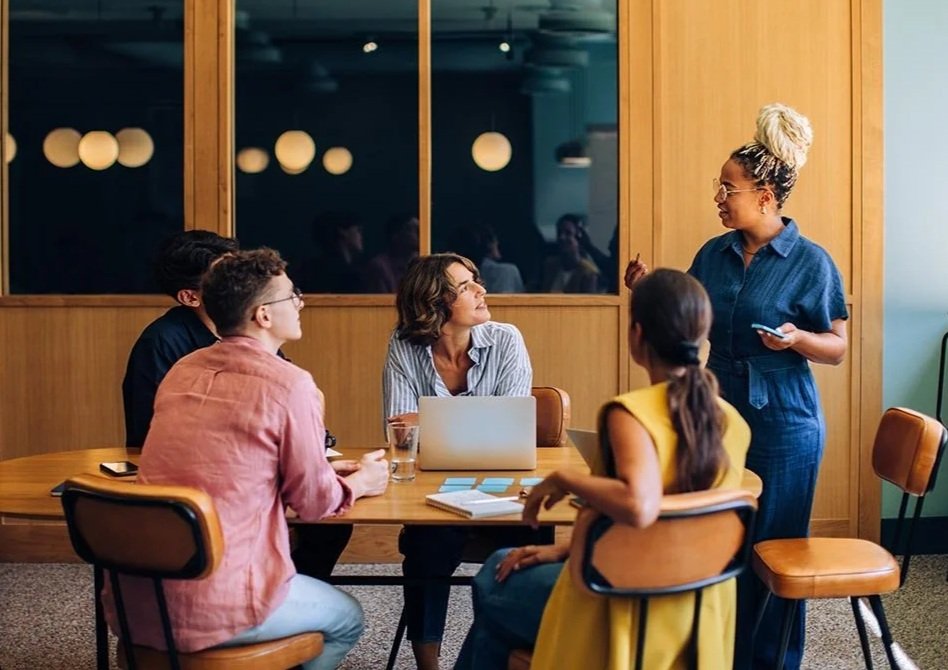 Woman talking to nonprofit team sitting along a long meeting table with sticky notes on it in a wood panel room.
