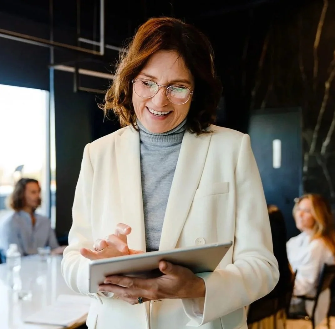 Woman wearing cream blazer looking at notes on tablet while standing near meeting table.j