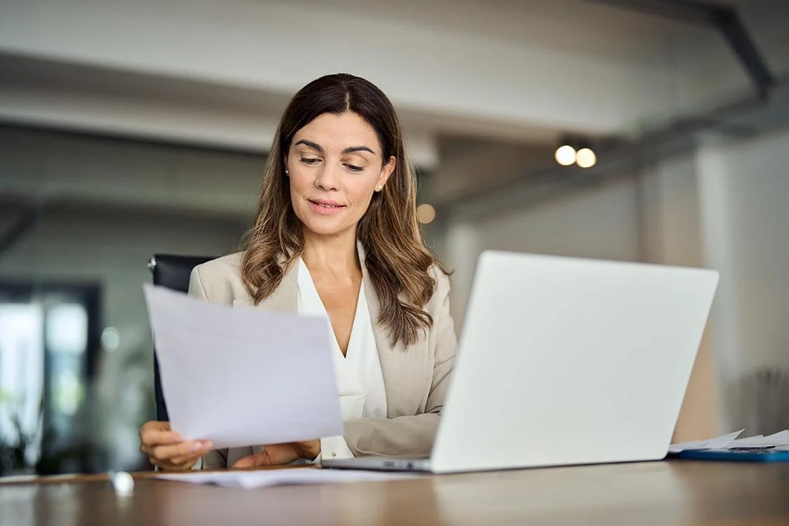 Woman in professional business attire carefully reviewing paperwork and working on her laptop.