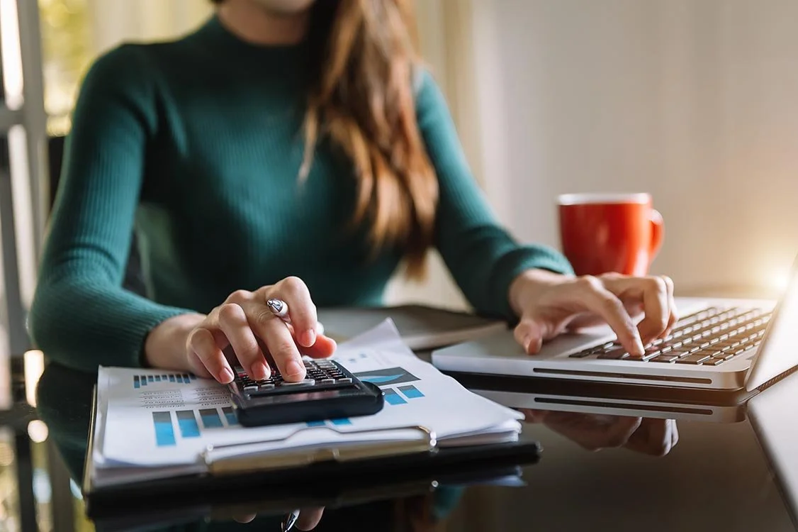 woman crunching numbers on a calculator with reports beneath it while typing on her laptop